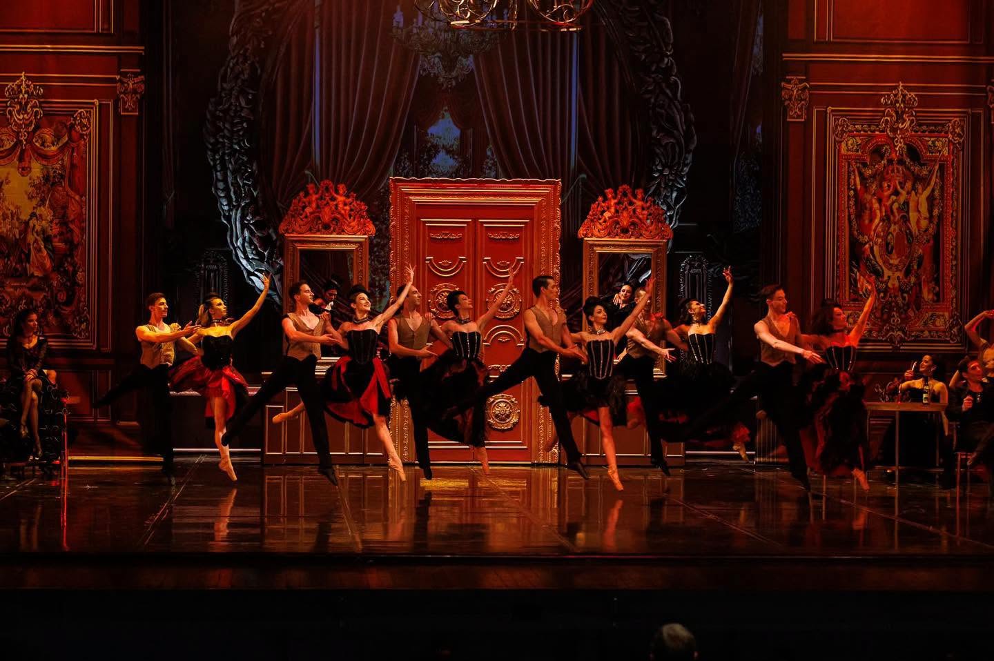 Ballet dancers performing a synchronized mid-air leap in pairs on an ornate stage set