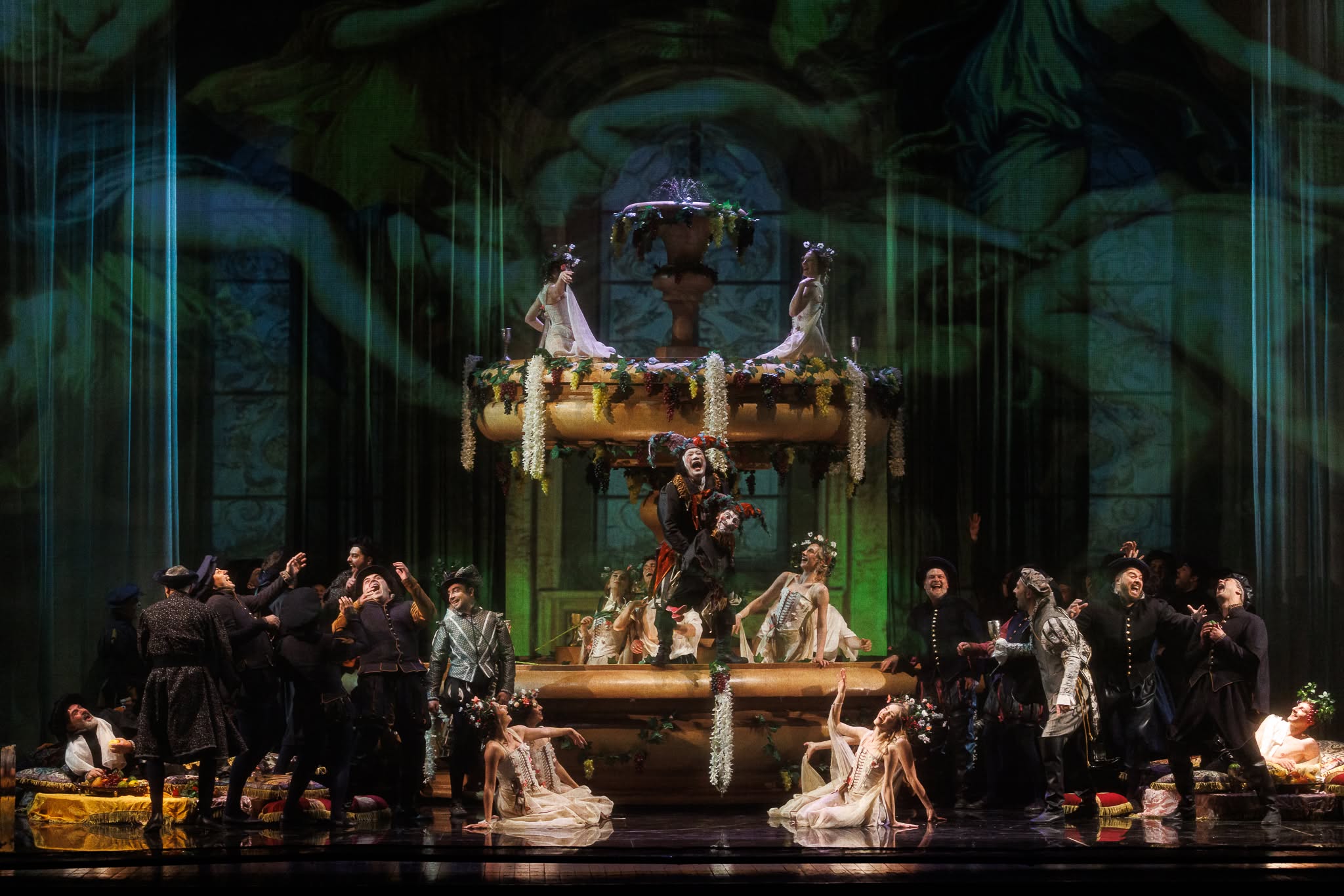 Opera performers in elaborate costumes gathered around a large ornate stone fountain set with performers posing on top and around its base