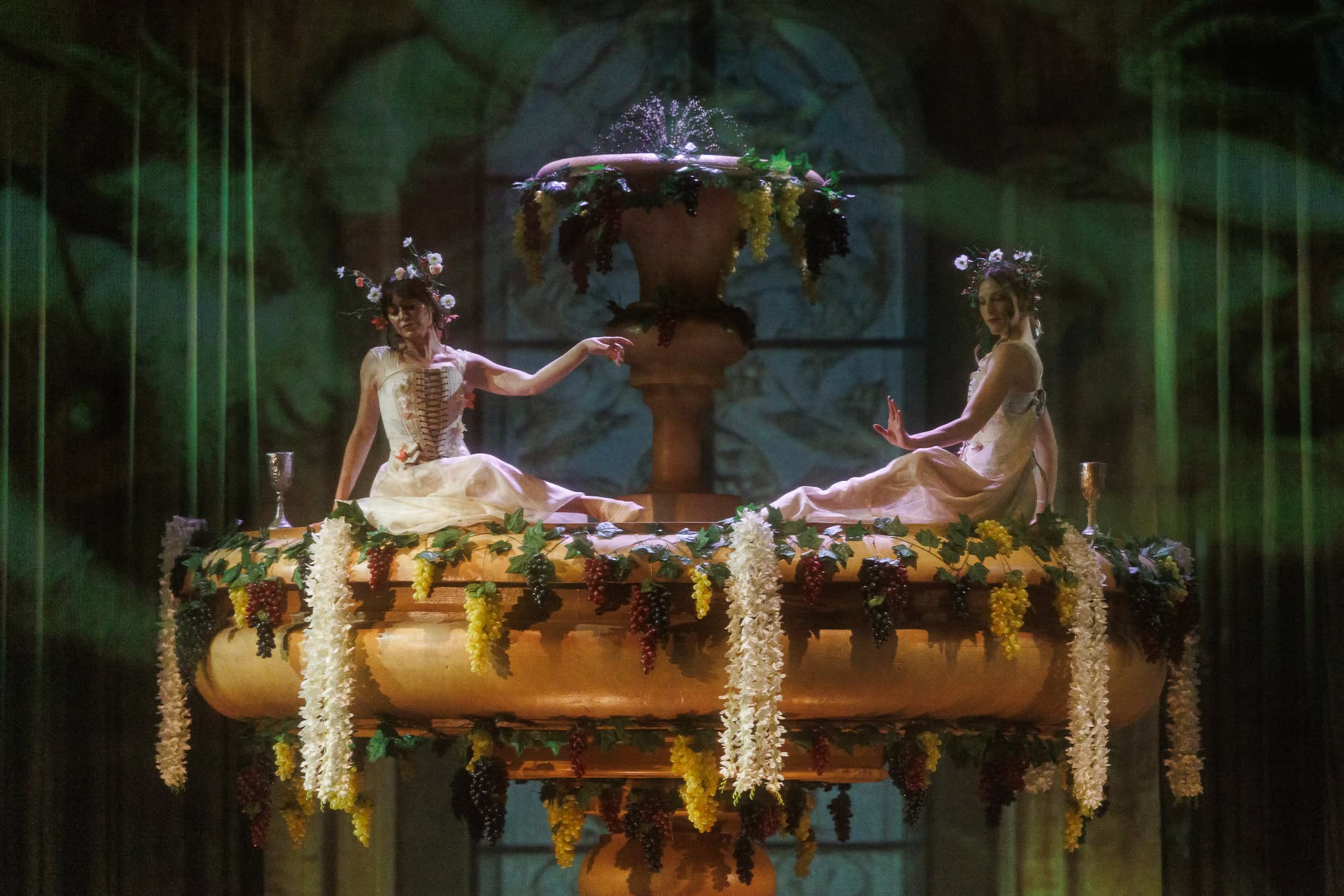 Two female performers in light dresses and floral hairpieces posing symmetrically atop a large fountain adorned with grapes and garlands