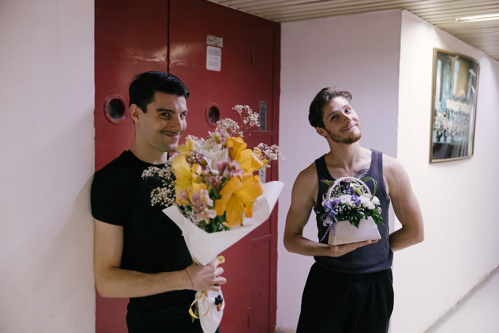 Two male performers smiling backstage in a hallway while holding floral bouquets
