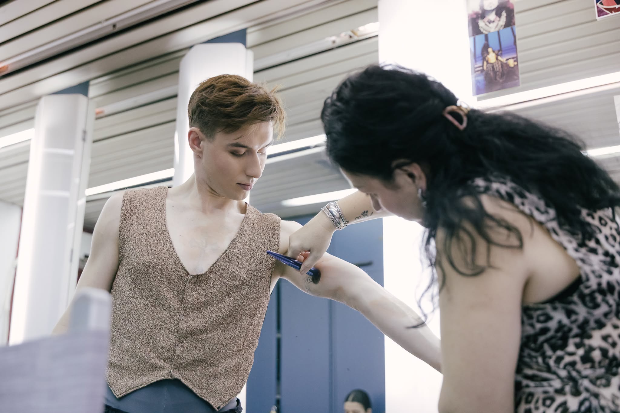 A makeup artist applying body makeup with a brush to a male ballet dancer's arm backstage