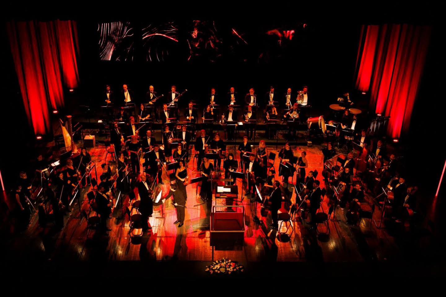 Full symphony orchestra performing on a stage under dramatic red lighting