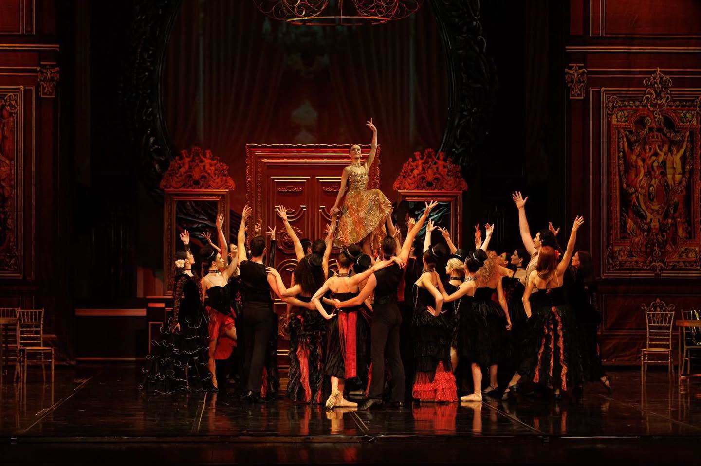 Group of ballet dancers in dark period costumes lifting a female soloist in a gold dress above them on stage