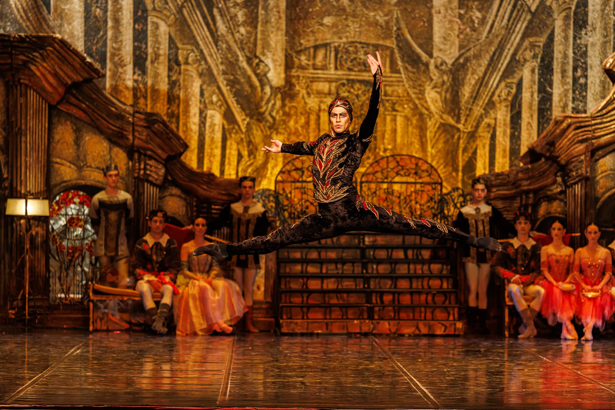 Male ballet dancer in a black and red patterned costume performing a high split leap in mid-air on stage