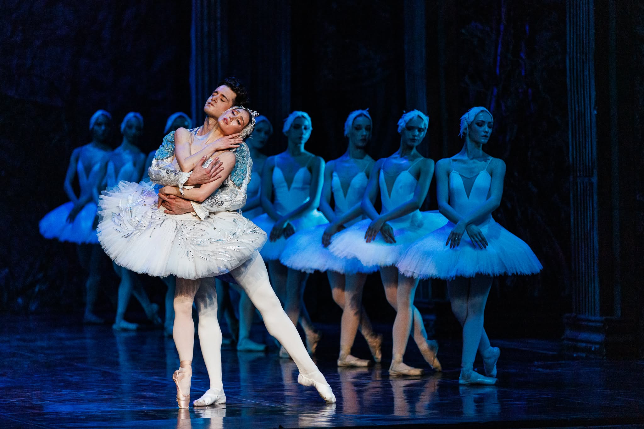 Male and female ballet lead dancers embracing in white costumes with a corps de ballet of swans in the background